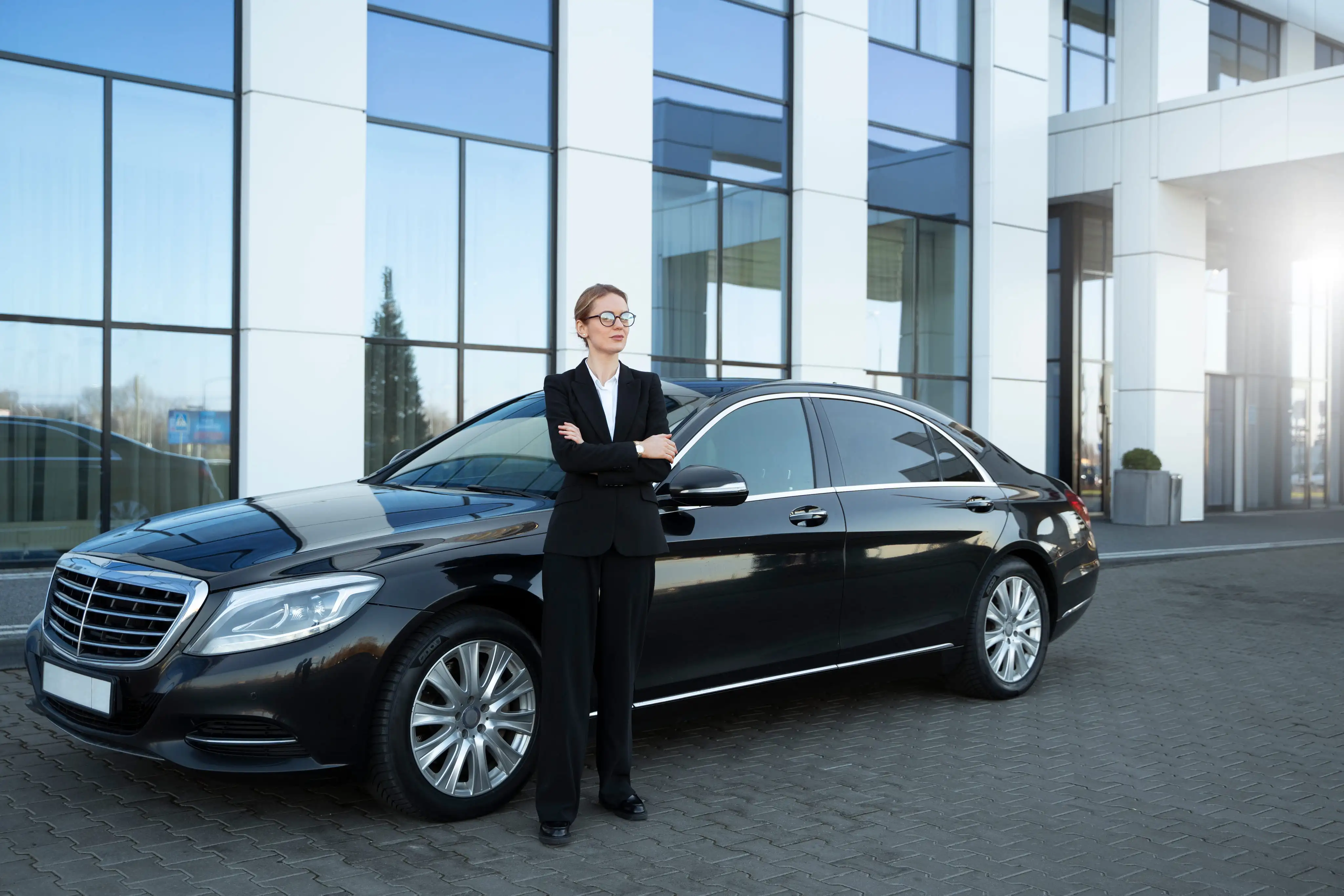 A chauffeur standing beside a black Mercedes sedan outside a glass office building